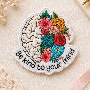 May include: Embroidered patch featuring a brain design on the left and colorful flowers on the right. The patch has the text "Be kind to your mind" in a curved font. The patch is white with a white border and is on a cream-colored fabric.