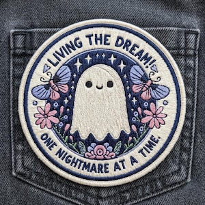May include: Embroidered patch with a cute ghost design, surrounded by butterflies, flowers, and stars. The patch has the text "LIVING THE DREAM! ONE NIGHTMARE AT A TIME." The patch is circular and has a navy blue and cream color scheme.