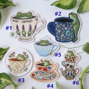 May include: Embroidered patches featuring frogs in teacups. Designs include a frog in a lavender-decorated teacup, a frog with a teabag, and a frog in a teacup with a spoon. The patches are colorful and detailed.