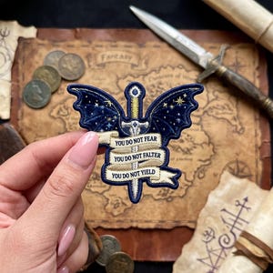 May include: Embroidered patch featuring a sword with dark blue wings and the text "YOU DO NOT FEAR, YOU DO NOT FALTER, YOU DO NOT YIELD." The patch has gold and white accents, with a starry night sky design on the wings. Fantasy theme.