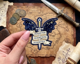 May include: Embroidered patch featuring a sword with dark blue wings and the text "YOU DO NOT FEAR, YOU DO NOT FALTER, YOU DO NOT YIELD." The patch has gold and white accents, with a starry night sky design on the wings. Fantasy theme.