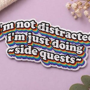 May include: Embroidered patch with the text "I'm not distracted I'm just doing side quests" in white, outlined in black, with a rainbow gradient. The patch has a white border and a curved shape.