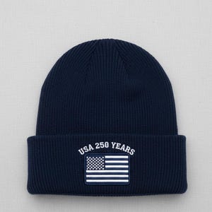 May include: A navy blue knitted beanie hat with a turned-up brim. It has a patch displaying the American flag and the text "USA 250 YEARS" in white. This is a classic style of hat, suitable for cold weather.