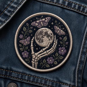 May include: Embroidered patch featuring a skeletal hand holding a detailed moon. The design includes purple butterflies, flowers, and stars on a dark blue background. The patch is circular and has a stitched border, suitable for jackets or bags.