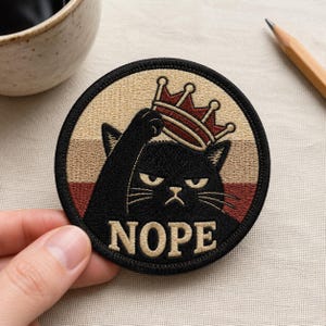 May include: Round patch featuring a black cat wearing a crown, with the word "NOPE" in beige. The cat has an annoyed expression. The patch has a beige, brown, and black color scheme.