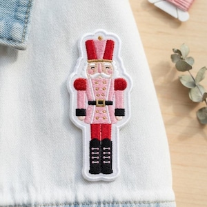 May include: An embroidered patch featuring a nutcracker figure with a pink jacket, red hat, red trousers, and black boots. The nutcracker has gold-colored buttons, a black belt with a gold buckle, and a white mustache, all outlined in white thread.