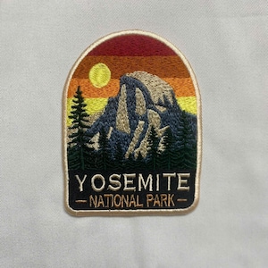 May include: An embroidered patch featuring a mountain landscape with a sunset sky in shades of red, orange, and yellow. A yellow sun, dark green pine trees, and a grey mountain peak are depicted. The text YOSEMITE NATIONAL PARK is in white.
