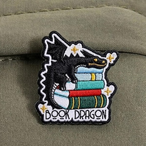 May include: Embroidered patch showing a black dragon on a pile of books. The patch has a white border and the words "BOOK DRAGON" are printed in white. The background is a muted green fabric.