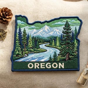 May include: A rectangular embroidered patch shaped like the state of Oregon, featuring a detailed landscape of snow-capped mountains, a winding blue river, and dense green pine forests. The word OREGON is stitched in white at the bottom.