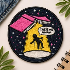 May include: Embroidered patch with a dark blue background, featuring a pink book emitting a yellow beam of light. A black silhouette is being lifted by the light, with the text "TAKE ME AWAY" in a speech bubble. White and pink stars surround the image.