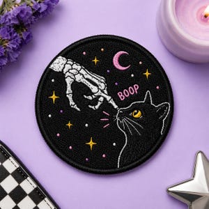 May include: A black, round patch with a skeleton hand booping a black cat's nose. The patch features a pink crescent moon, stars, and the word "BOOP". The design is embroidered with white, yellow, and pink thread.