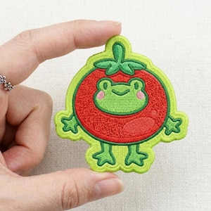 May include: Embroidered patch featuring a cartoon frog inside a red tomato. The frog is green with pink cheeks, and the tomato has a green outline. The patch is held up by a hand.