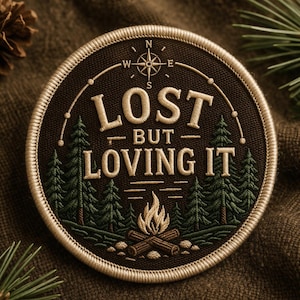 May include: Round embroidered patch with the text "LOST BUT LOVING IT" in cream-colored thread. The patch features a compass rose, pine trees, and a campfire design on a dark brown background, with a cream-colored border.