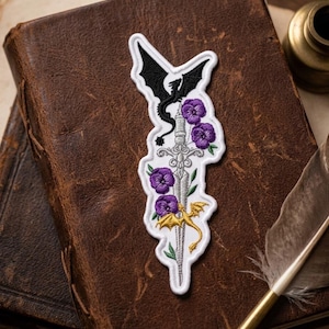 May include: Embroidered patch featuring a sword, dragons, and purple flowers. The patch has a white border and includes a black dragon at the top and a gold dragon at the bottom. The design is set against a brown leather background.