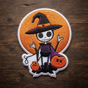 May include: A circular embroidered patch featuring a white skeleton character wearing a black witch hat with an orange band. The skeleton stands before a large orange moon, flanked by two small orange pumpkins, with a purple cape and white accents.