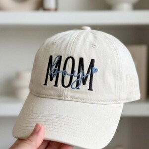 May include: Cream-colored baseball cap with the word "MOM" embroidered in black block letters, with "boy" in light blue script underneath. The cap has a curved brim and a classic six-panel design.