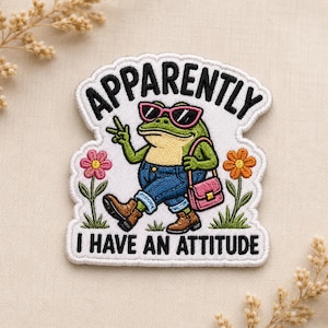 May include: An embroidered patch featuring a green frog wearing pink sunglasses, blue jeans, and brown boots, carrying a pink handbag. The frog is making a peace sign. The text reads APPARENTLY I HAVE AN ATTITUDE. Two flowers flank the frog.