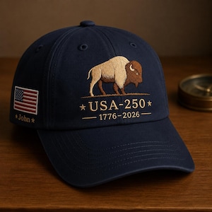 May include: A navy blue baseball cap featuring an embroidered brown and tan bison above the text USA-250 and 1776-2026. The side of the cap displays an embroidered American flag patch with the name John written in gold thread below the flag.