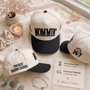 May include: Cream-colored baseball caps with black visors. One cap has "MOMMIN'" embroidered across the front, another says "TOO BUSY RAISING LEGENDS", and a third displays "24/7". The caps are part of "The Mommin' Collection".