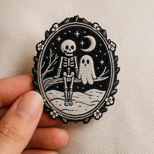 May include: Oval-shaped embroidered patch featuring a skeleton and a ghost under a crescent moon and stars. The design is set against a black background, framed by a border of leaves and flowers. The patch is held by a hand.