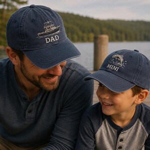 May include: Two navy blue baseball caps. One cap features an embroidered boat design with the word "DAD". The other cap has a leaping fish design with the word "MINI". Both caps are worn by a father and child.