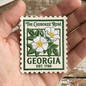 May include: A decorative stamp featuring the Cherokee Rose, the state flower of Georgia. The stamp has a cream background with a green border and text. The image shows two white flowers with yellow centers and green leaves. The text reads "The Cherokee Rose" and "Georgia Est. 1788."