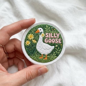 May include: Embroidered patch featuring a white goose with a flower garland around its neck, surrounded by green grass and yellow flowers. The words "SILLY GOOSE" are embroidered in pink. The patch has a white border.