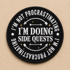 May include: Black circular patch with white text that reads "I'M NOT PROCRASTINATING I'M DOING SIDE QUESTS". The patch has a black border and white arrows and stars. The background is a tan fabric.