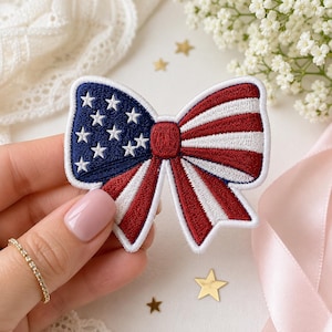 May include: Embroidered bow patch in red, white, and blue, featuring a patriotic design with stars and stripes. The bow has a white border and is held by a hand. Perfect for adding a touch of Americana to clothing or accessories.