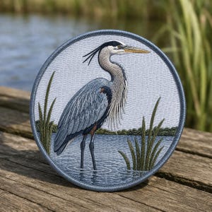 May include: Embroidered patch featuring a detailed illustration of a Great Blue Heron standing in water. The patch is circular, with a blue border and a light blue background. The heron is depicted in shades of blue, gray, and brown, with long, slender legs and a long beak.