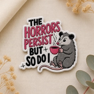 May include: Embroidered patch featuring a gray opossum holding a pink mug, with the text "THE HORRORS PERSIST BUT SO DO I" in black and pink. The patch has a white border and is on a neutral background.