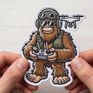 May include: Embroidered patch depicting a cartoon Bigfoot character in aviator goggles and a vest, holding a drone remote control. The patch is brown, green, and white, with a drone above. The patch is held by two hands.