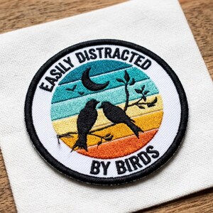 May include: Embroidered patch with the text "EASILY DISTRACTED BY BIRDS." The patch features a circular design with a black outline, a crescent moon, two birds, and a colorful striped background in shades of blue, yellow, and orange.