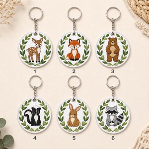 May include: Six circular keychains featuring embroidered woodland animals: a fawn, fox, bear, skunk, rabbit, and raccoon. Each animal is centered within a green leaf wreath on a white background, attached to a silver metal ring and chain.