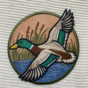 May include: A circular embroidered patch featuring a mallard duck in flight over a blue water surface. The background depicts a tan sky with green cattails and reeds. The duck has a green head, yellow beak, and white and green wing details.