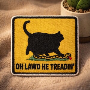 May include: Embroidered patch with a black cat silhouette stepping on a snake, set against a yellow background. The patch has the text "OH LAWD HE TREADIN'" in white. The patch is square with a black border.