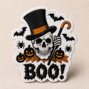 May include: An embroidered patch featuring a white skull wearing a black top hat with an orange band, holding a cane. The design includes two orange jack-o'-lanterns, brown leaves, black spiders, and bats, with the word BOO! in black text.