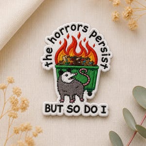 May include: Embroidered patch featuring a cartoon opossum standing in front of a green dumpster on fire. The patch reads "the horrors persist" above the dumpster and "but so do I" below. The patch has a white border.