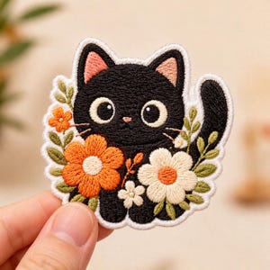 May include: Embroidered patch featuring a black cat with large, round eyes and pink inner ears. The cat is surrounded by orange and white flowers with green leaves. The patch is held by a hand.