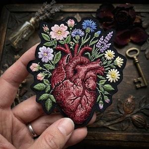 May include: An embroidered patch on a black fabric base featuring a detailed anatomical heart in shades of deep red and burgundy, surrounded by a vibrant arrangement of colorful wildflowers including pink roses, blue cornflowers, and yellow daisies.