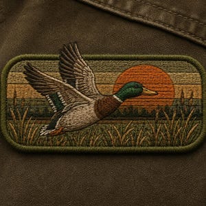 May include: Embroidered patch featuring a mallard duck in flight against a sunset backdrop. The patch has a rectangular shape with a green border. The duck has brown, white, and green feathers. The sunset is orange and yellow.