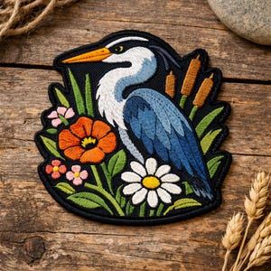 May include: Embroidered patch featuring a blue heron with white and blue feathers, an orange beak, and a black outline. The patch includes orange, pink, and white flowers, green leaves, and cattails against a black background.