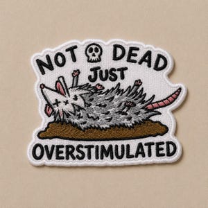 May include: Embroidered patch featuring a cartoon opossum lying on its back, appearing to play dead. The patch has the text "NOT DEAD JUST OVERSTIMULATED" in black lettering with a skull graphic. The opossum is gray with pink ears and a pink tail.