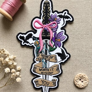 May include: Embroidered patch featuring a sword design with purple flowers, a pink bow, and the text "None of this is worth it without you." The patch has a black border and is on a tan background. A spool of pink thread and a wooden button are also visible.