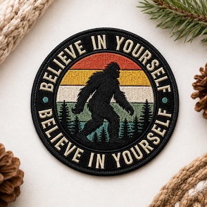 May include: Circular black embroidered patch with a Bigfoot silhouette walking across a forest. The patch features the words "BELIEVE IN YOURSELF" in a circular design, set against a retro sunset background.