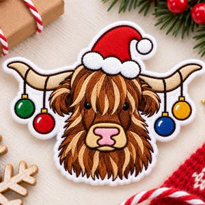 May include: An embroidered patch featuring a brown Highland cow wearing a red and white Santa hat. The cow has long horns decorated with four hanging Christmas ornaments in green, red, blue, and yellow. The patch has a white stitched border.