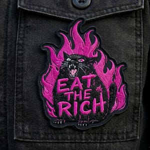 May include: Embroidered patch showing a black cat in pink flames, with the text "EAT THE RICH". The patch is attached to a black denim jacket. The cat is snarling, with its mouth open.