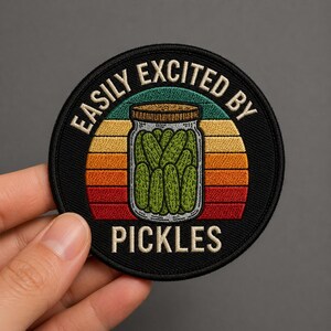 May include: Round black patch with the text "EASILY EXCITED BY PICKLES" in white. The patch features a jar of green pickles in the center, set against a sunset design with orange, yellow, and green stripes.