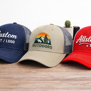 May include: Three baseball caps with mesh backs on a wooden surface. The caps are navy, beige, and red. The navy cap has the text "Custom TEXT / LOGO" in white. The beige cap has an outdoors graphic. The red cap has the text "Allstars".
