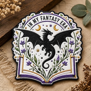 May include: Embroidered patch featuring a black dragon with outstretched wings, surrounded by floral elements and the text "IN MY FANTASY ERA". The design is set against a white background with purple and gold accents, resembling an open book.